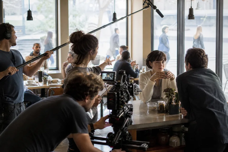 Ekipa filmowa nagrywa reklamę w kawiarni, w tle statyści przy stolikach, asystentka reżysera z listą ustawia tło